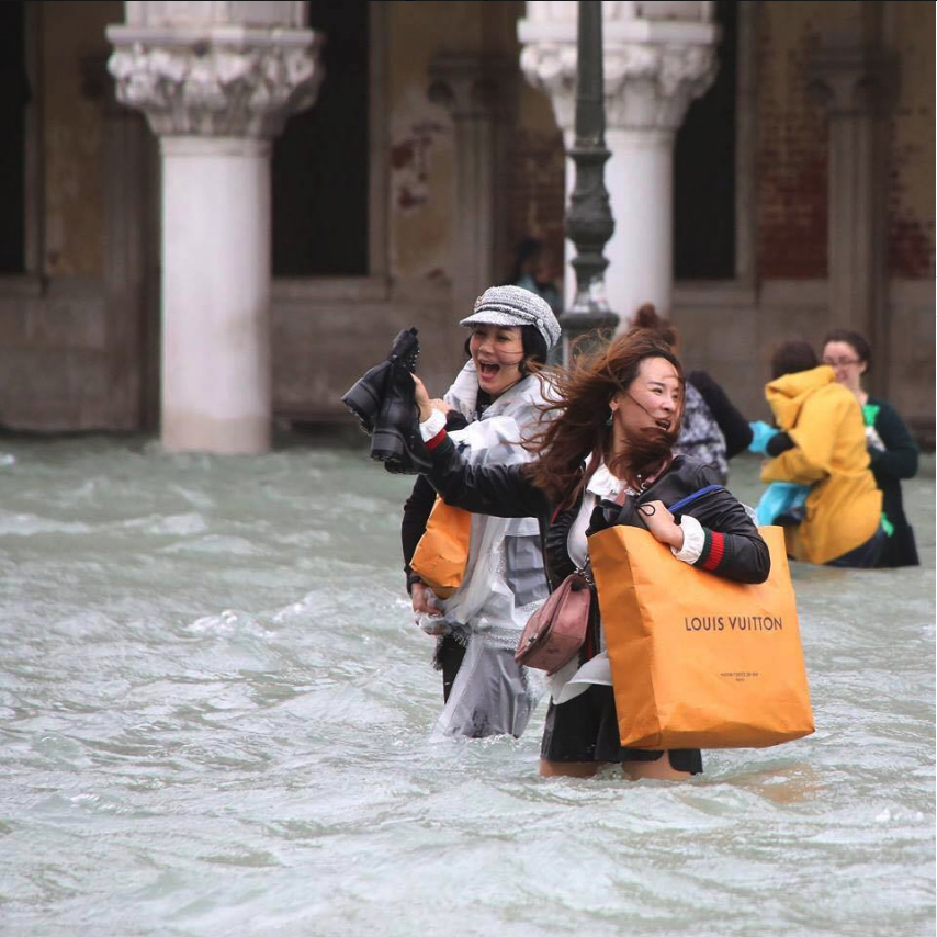 Shopping while they drawn. | Tourist with Louis Vuitton bag wading through flooded Venice.